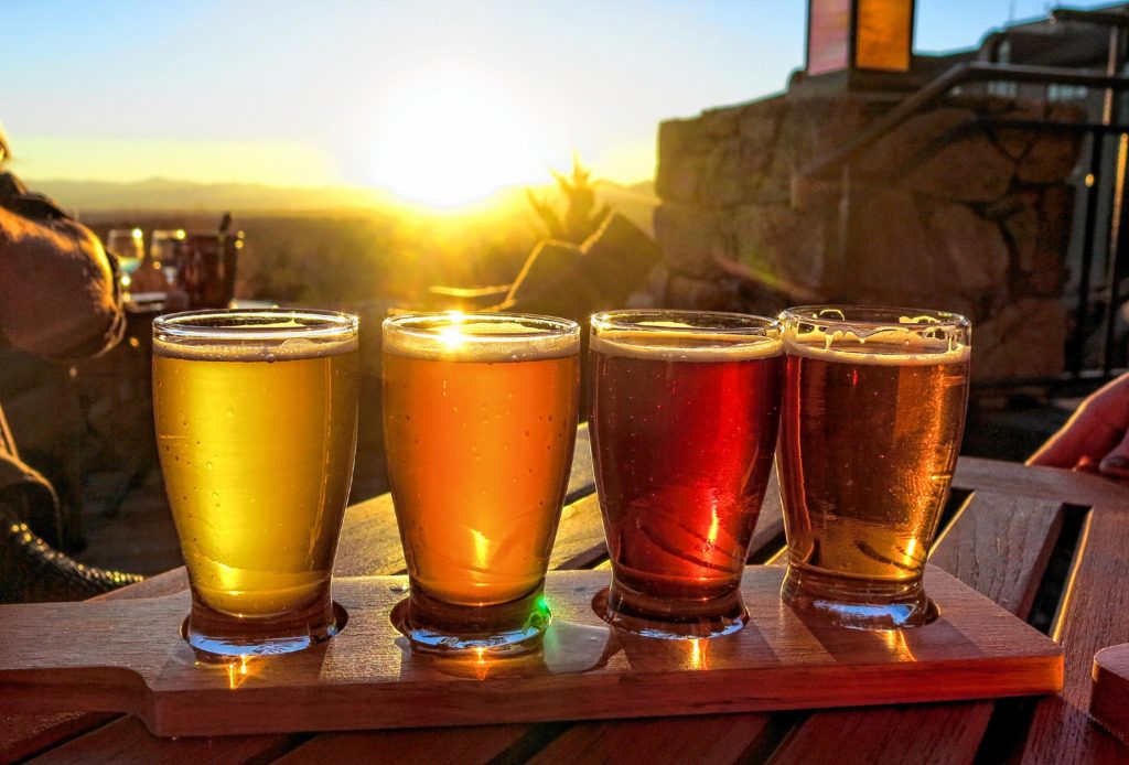 A colorful beer flights is illuminated against a bright sunset.  4 glasses of different beers are set on a wooden board and table outside.
