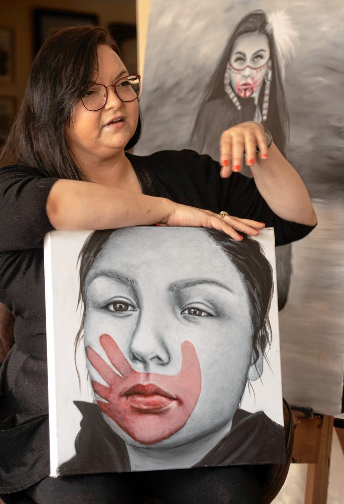 Nayana LaFond with two of the 77 MMIW portraits she’s painted. She holds a painting of Kaysara, a young Native American woman from Montana who was murdered; behind her is one of Littlefox Steeprock, a survivor of violence and now an advocate for MMIW.