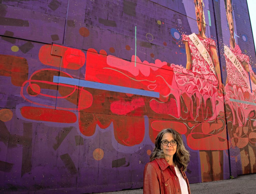 Britt Ruhe, the director of  Commonwealth Murals in Springfield, stands in front of a mural called Victoria painted by Andrae and Priya Green on Taylor Street,  one of the 36 murals in town.